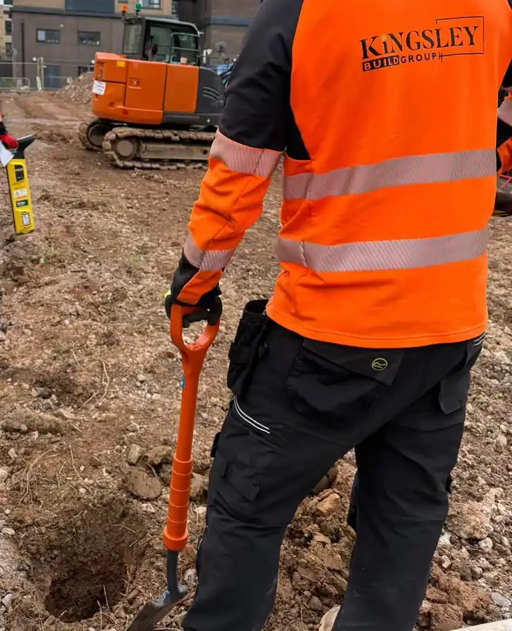Kingsley operative with a spade at a post hole, cable-avoidance scanner and excavator behind, on a groundworks site